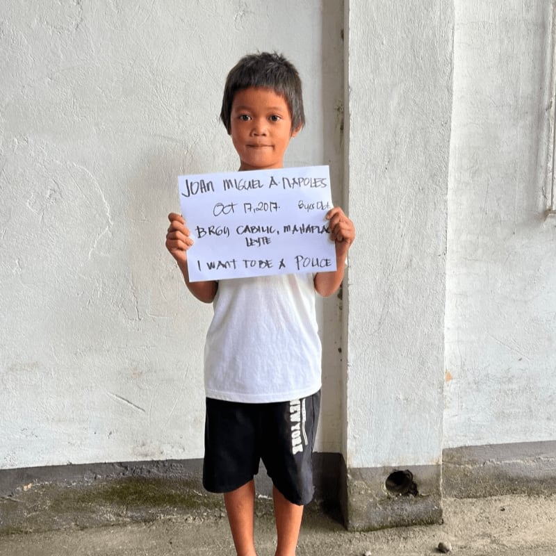 A young boy in a white t-shirt and black shorts holds a handwritten sign while standing against a plain white wall.