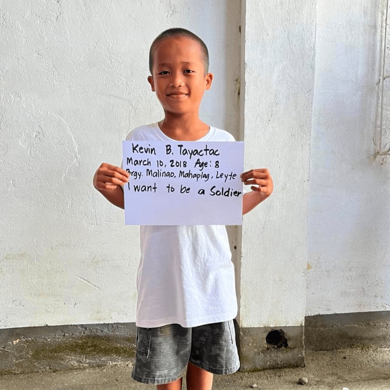 A young boy in a white t-shirt and grey shorts holds a handwritten sign against a plain white wall.
