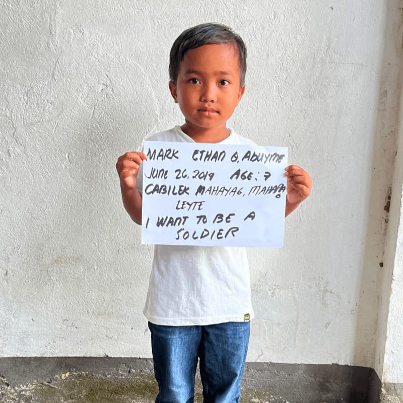 A young boy in a white t-shirt and blue jeans holds a handwritten sign against a plain, textured white wall.