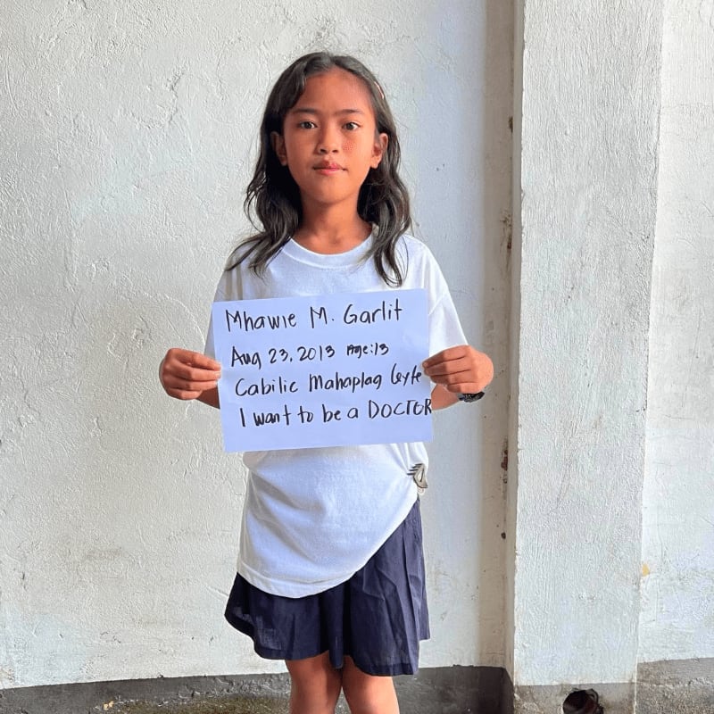 A young girl in a white t-shirt and dark skirt holds a handwritten sign against a plain, textured white wall.