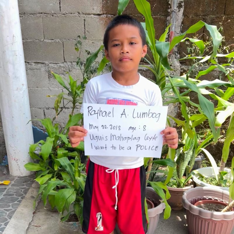 A young boy holds a handwritten sign in the foreground, standing before a backdrop of green plants and a grey cinder block wall.