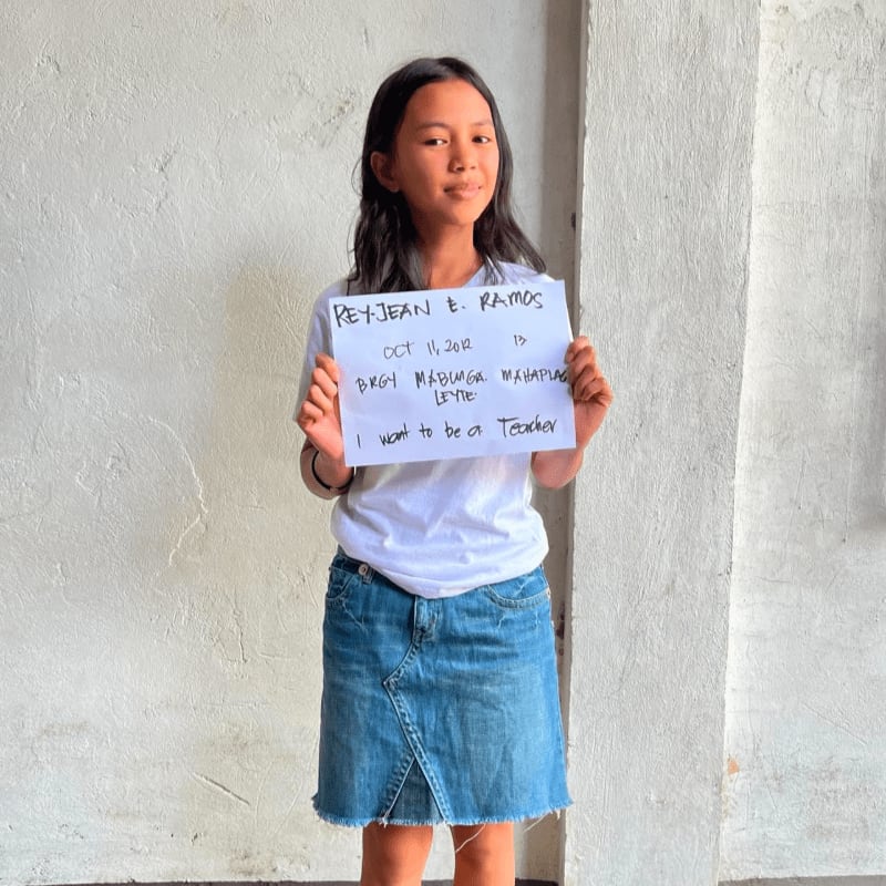 A young girl in a white t-shirt and denim skirt holds a handwritten sign while standing against a plain, light-colored wall.