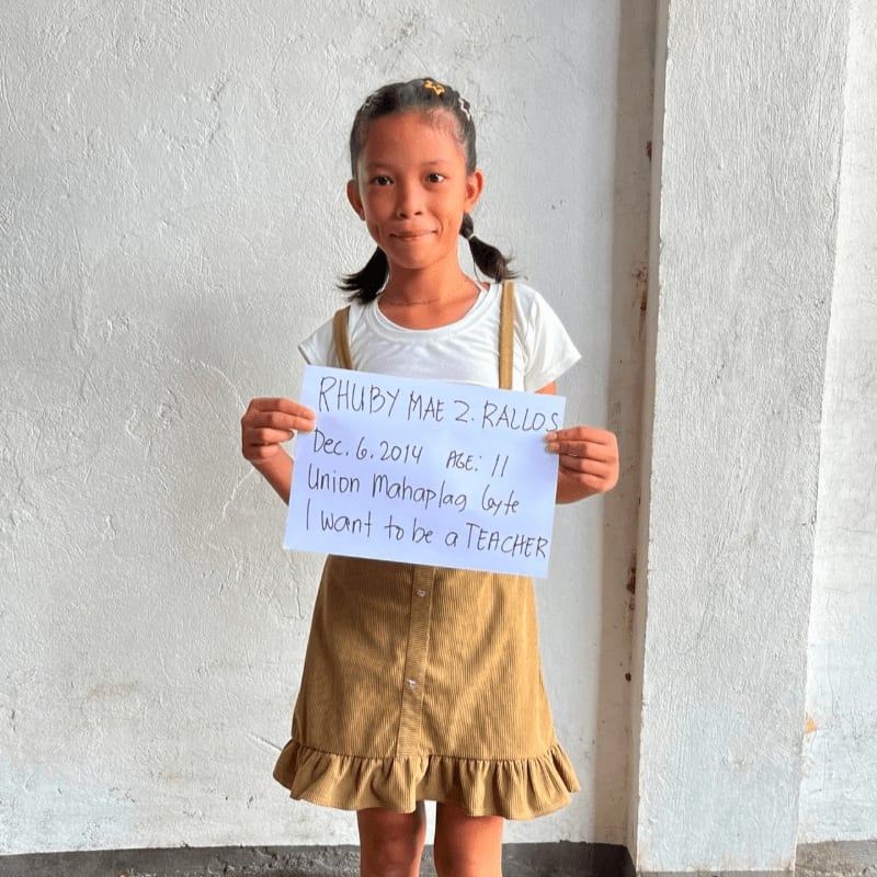 A young girl in a brown corduroy dress holds a handwritten sign against a plain white wall.