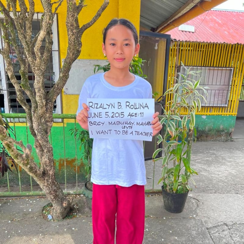A young girl in a white shirt and red pants holds a sign in the foreground, standing before a yellow building and green foliage in the background.