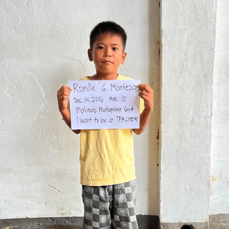 A young boy in a yellow shirt and checkered shorts holds a handwritten sign in front of a plain white wall.