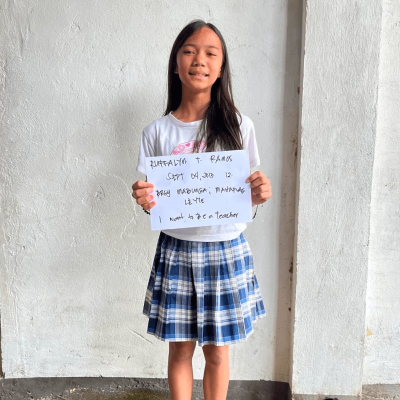 A young girl in a white t-shirt and blue plaid skirt holds a handwritten sign while standing against a plain white wall.