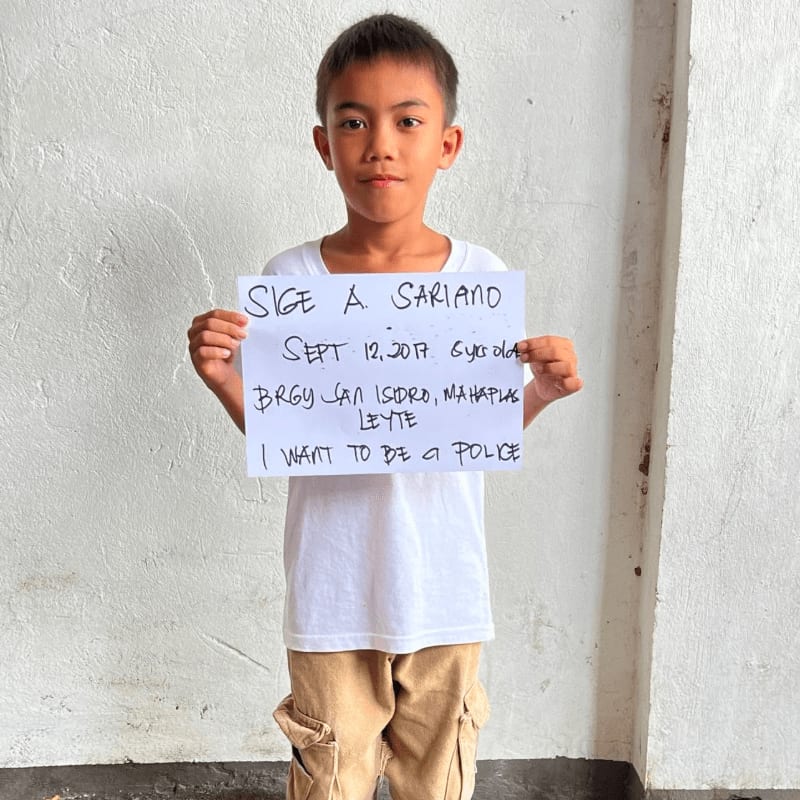 A young boy in a white t-shirt and khaki pants holds a handwritten sign in front of a plain white wall.