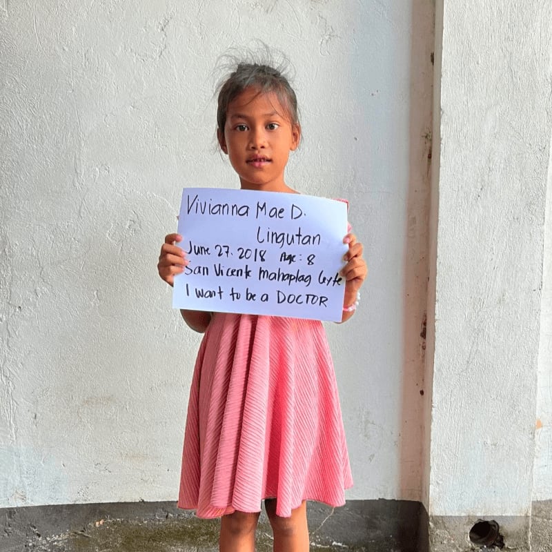 A young girl in a pink dress holds a handwritten sign in front of a plain white wall.