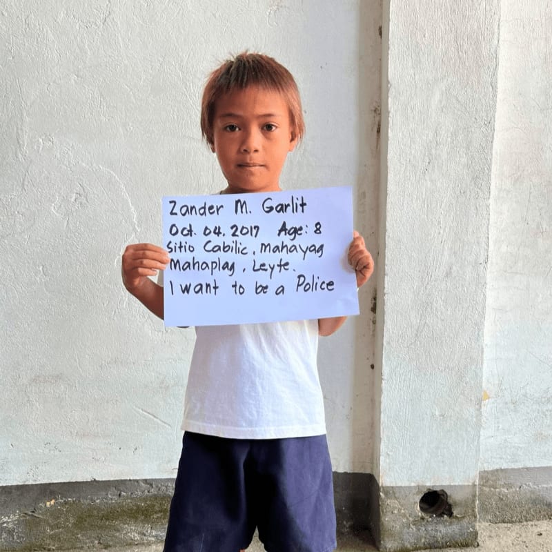 A young boy holds a handwritten sign against a plain, light-colored wall.