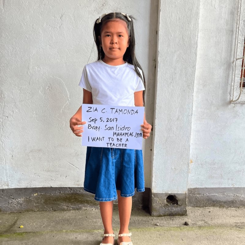 A young girl wearing a white t-shirt and blue skirt holds a sign stating she wants to be a teacher while standing against a plain white wall.