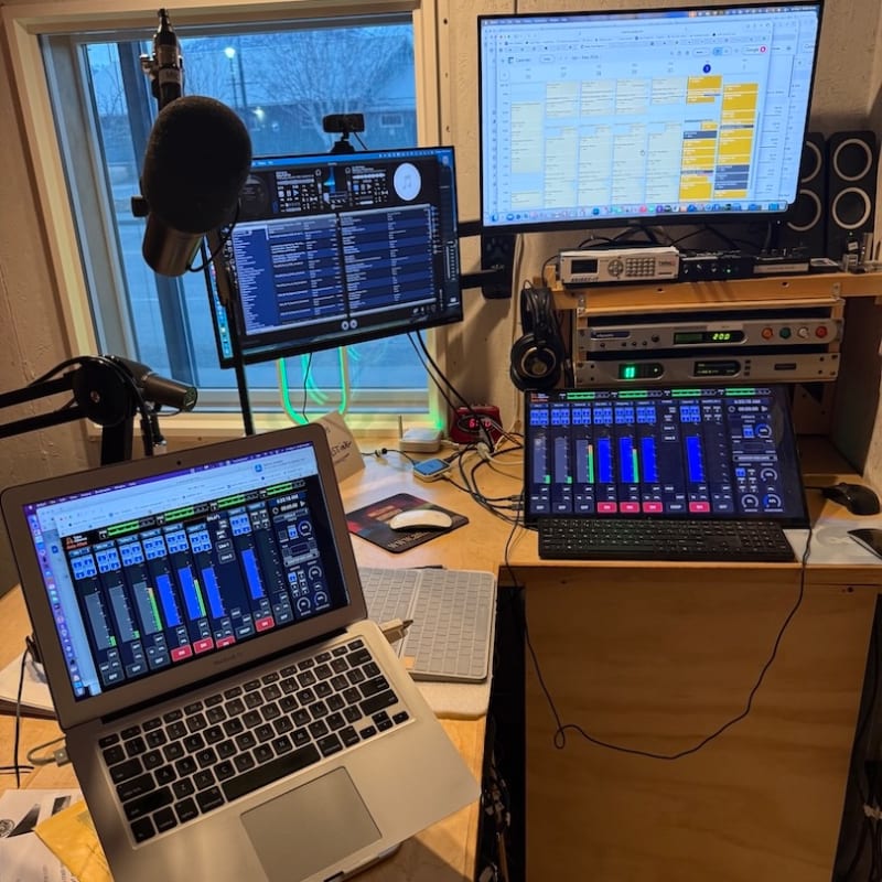 A laptop and audio equipment sit in the foreground of a home studio with multiple monitors and a window in the background.