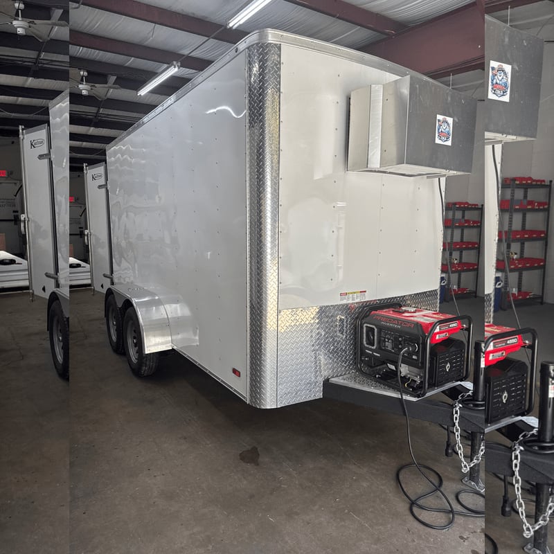 A silver enclosed utility trailer with a red generator on the tongue sits inside a warehouse with shelving in the background.