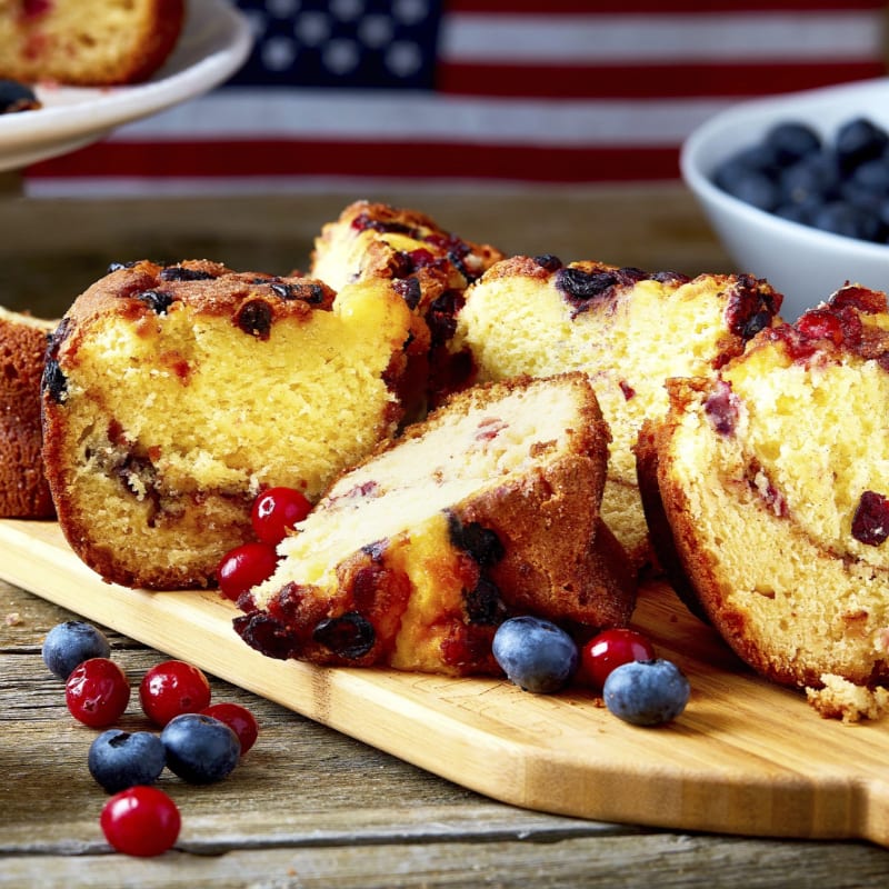 Slices of berry bread and fresh berries sit on a wooden cutting board with a blurred American flag in the background.