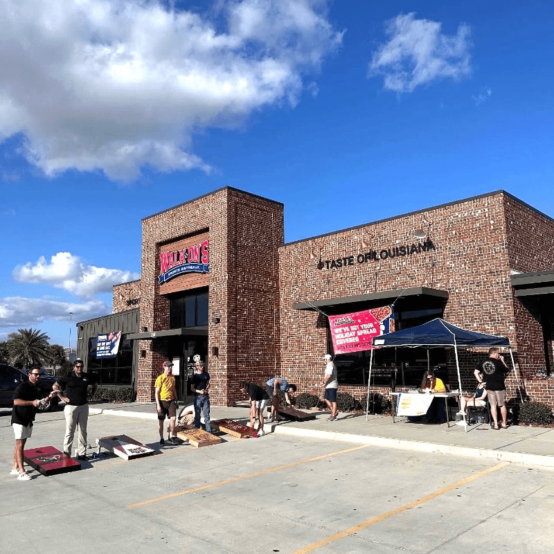 People gather in a parking lot in front of a brick building labeled "Taste of Louisiana" under a bright blue sky with fluffy clouds.