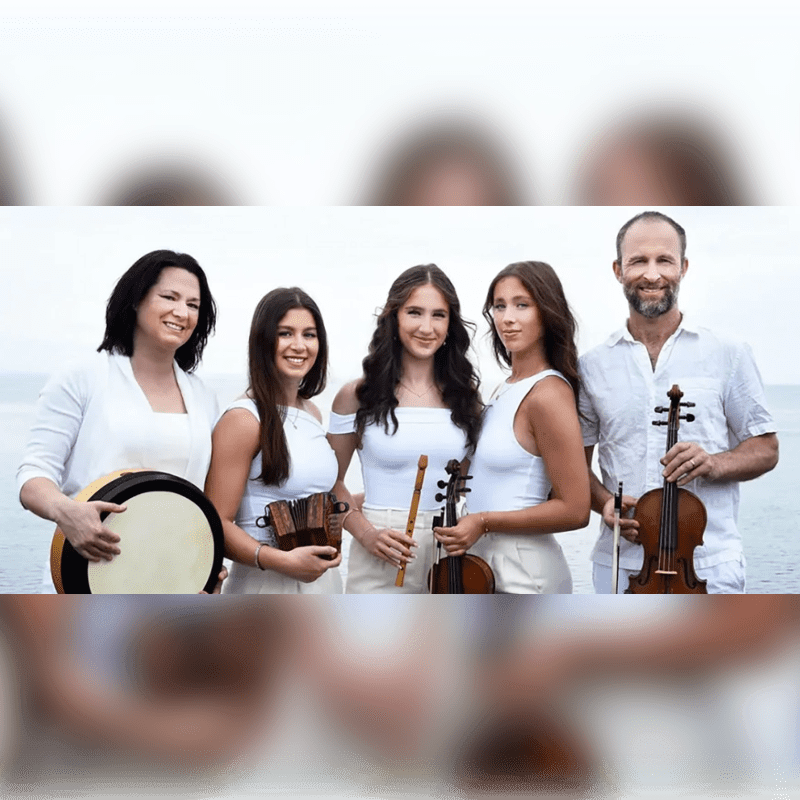 Five musicians dressed in white hold various instruments while posing together against a blurred coastal background.