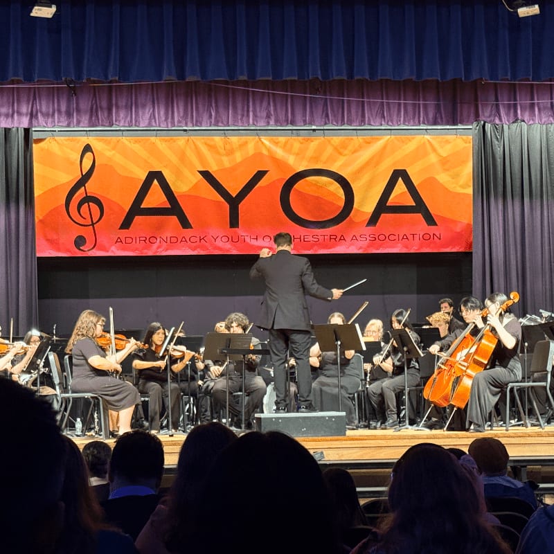A conductor leads a youth orchestra on a stage in front of a large orange "AYOA" banner, with an audience visible in the foreground.