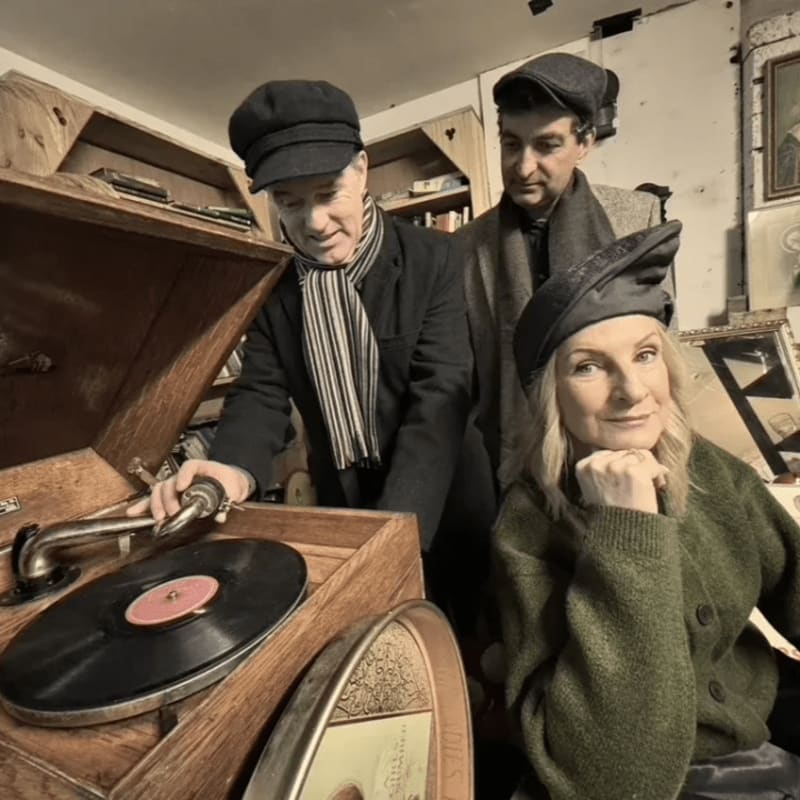 Three people in vintage attire gather around an old gramophone inside a cluttered room filled with wooden shelves and antiques.