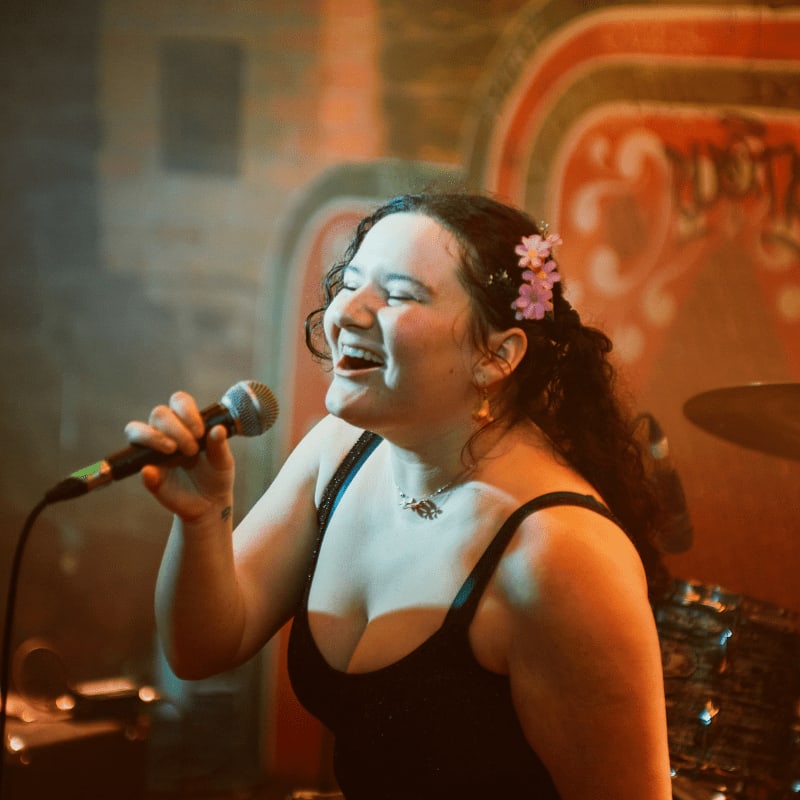 A woman with flowers in her hair sings into a microphone in the foreground, set against a blurred, warmly lit stage background.