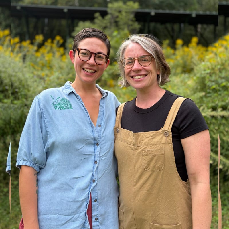 Two smiling women stand side-by-side in the foreground against a lush background of green foliage and yellow flowers.