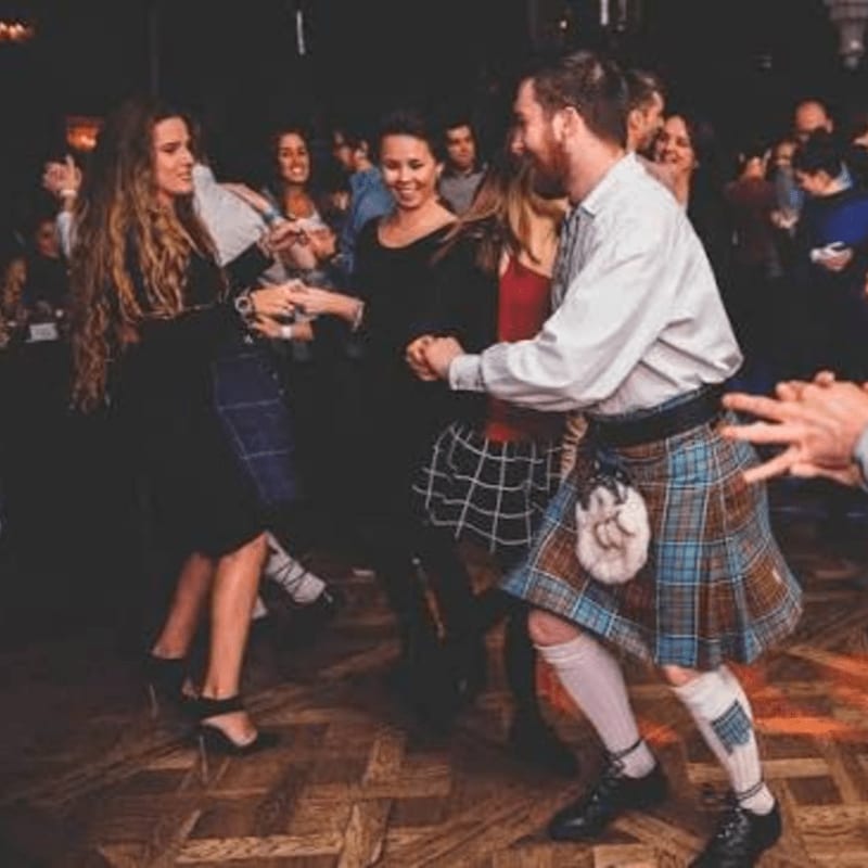People dance in a dimly lit room, with a man in a kilt and a woman in a black dress in the foreground and other guests blurred in the background.