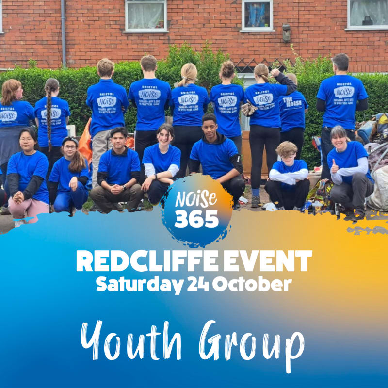 A group of young people in blue shirts pose together in the foreground against a background of a brick house and garden.