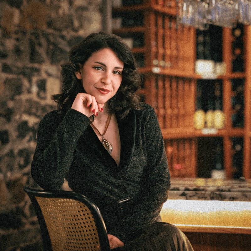 A woman in a dark blazer rests her chin on her hand in the foreground, with a stone wall and wine racks in the background.