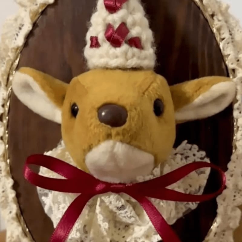 A plush deer wearing a white crocheted hat and a red bow sits against a dark wooden background framed by white lace.