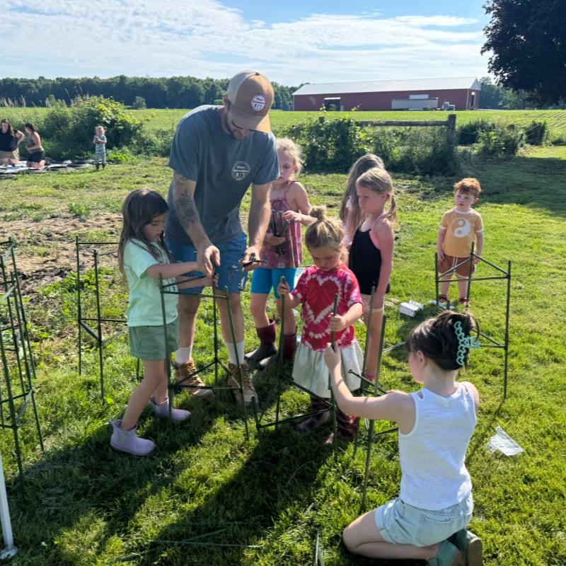 A man and several young children gather around green garden trellises in a grassy field with a red barn in the distant background.