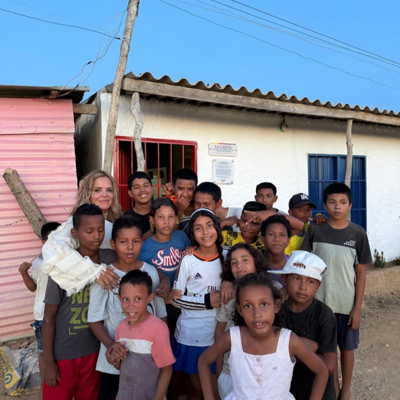 A group of children and a woman pose together in the foreground in front of a small white building with a pink wall and blue doors.