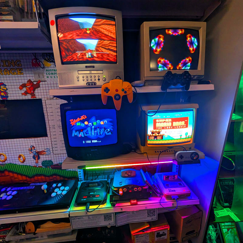 Multiple retro televisions and gaming consoles are arranged on white shelves against a wall decorated with posters and collectibles.