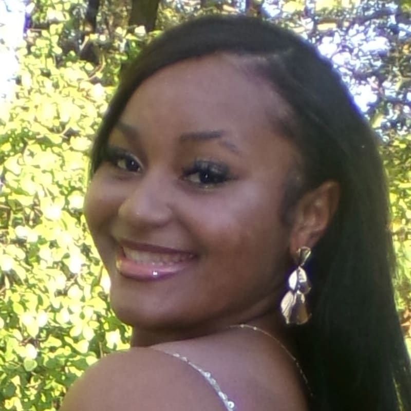 A smiling woman with long dark hair and gold earrings stands in the foreground against a background of lush green foliage.