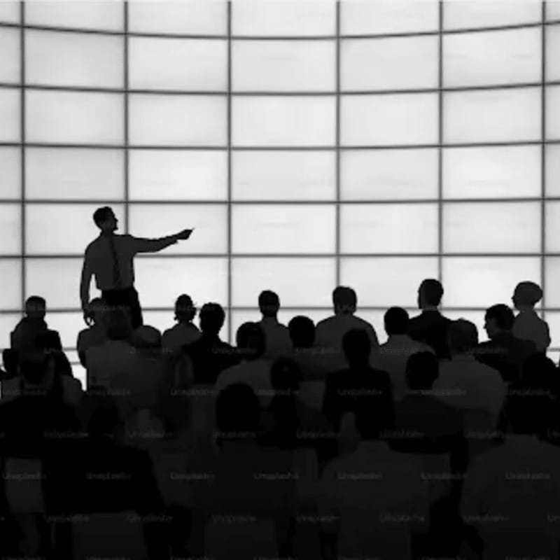 A man stands gesturing toward a large, gridded screen in the background while facing a silhouetted audience in the foreground.