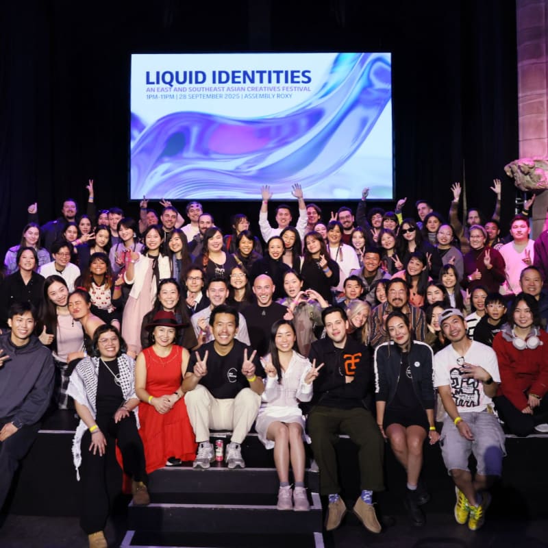 A large group of people pose for a photo in the foreground, while a bright screen displaying "LIQUID IDENTITIES" glows in the dark background.