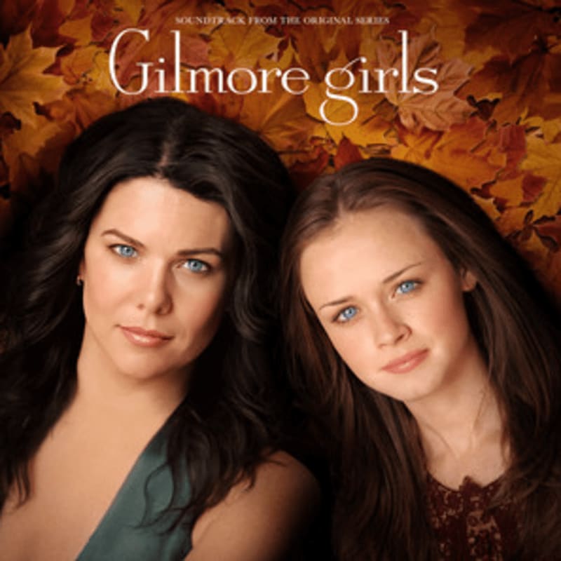 Two women with blue eyes pose closely together in the foreground against a background of autumn leaves and the text "Gilmore Girls."