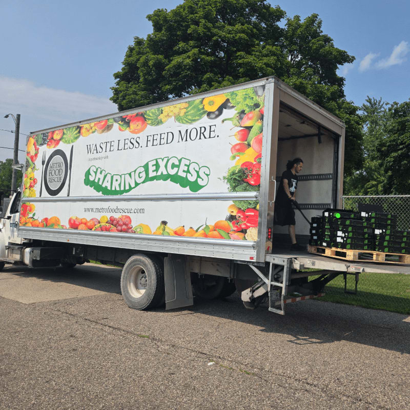 A woman loads crates of produce into a "Metro Food Rescue" truck parked on a paved lot with green trees in the background.