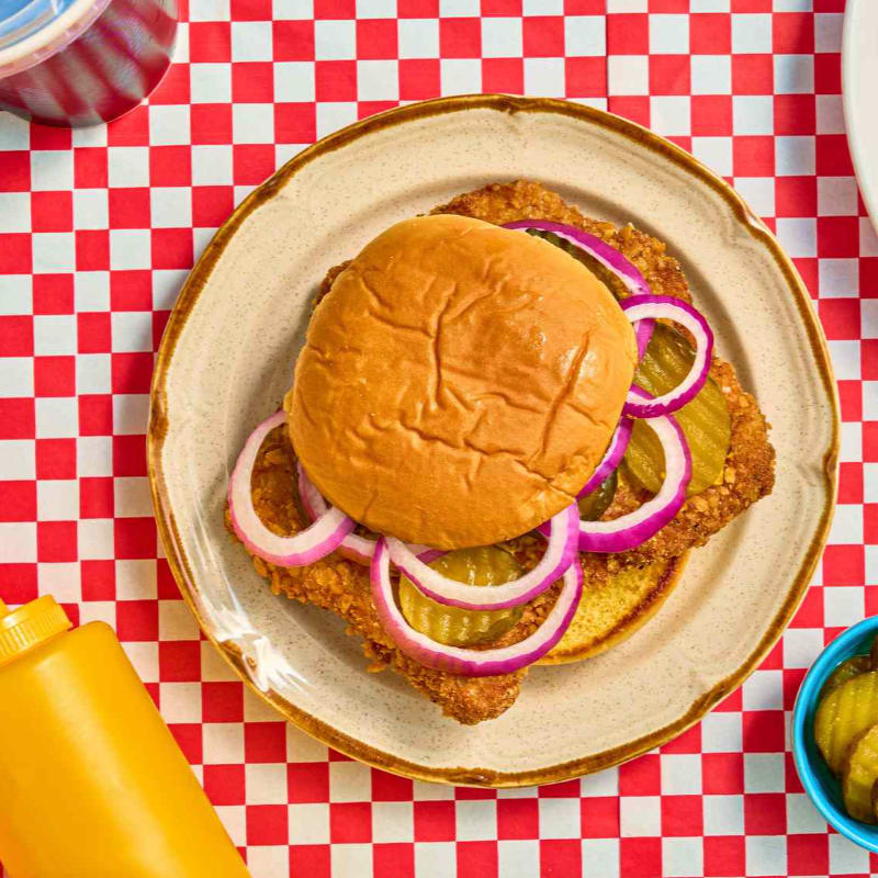 A fried chicken sandwich with red onions and pickles sits on a plate atop a red and white checkered tablecloth.
