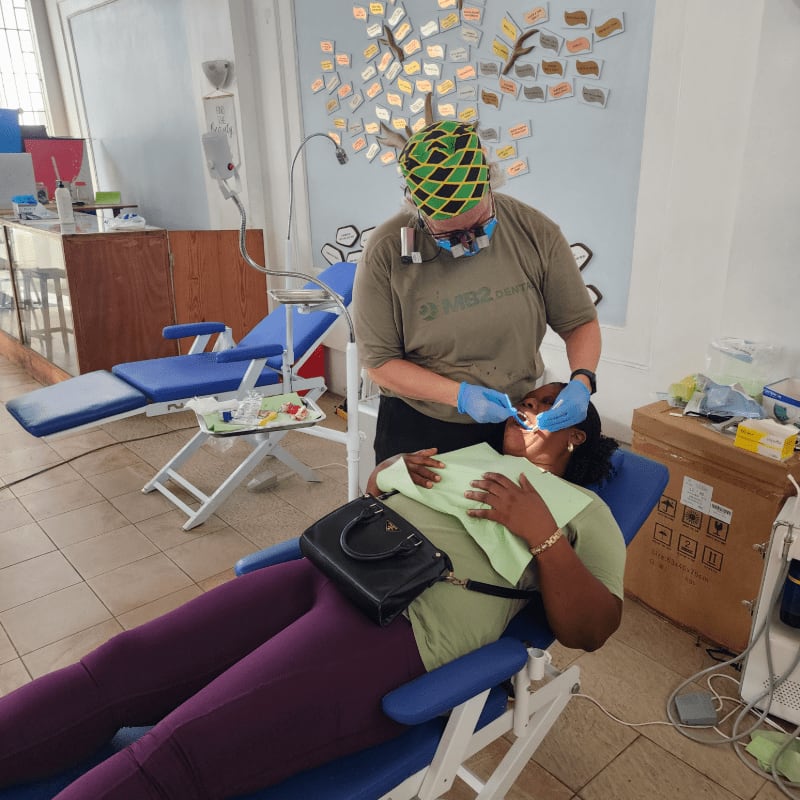 A dentist wearing a green patterned cap and mask treats a patient reclining in a blue dental chair within a clinic featuring white walls and medical equipment.