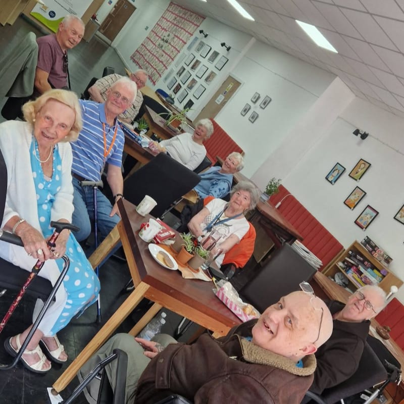 A group of elderly people gather around wooden tables in a brightly lit room decorated with framed pictures on the walls.
