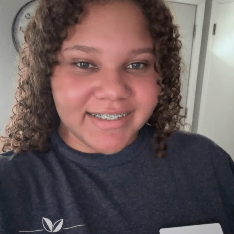 A smiling woman with curly hair and braces wears a dark blue shirt in front of a plain wall and a wall clock.