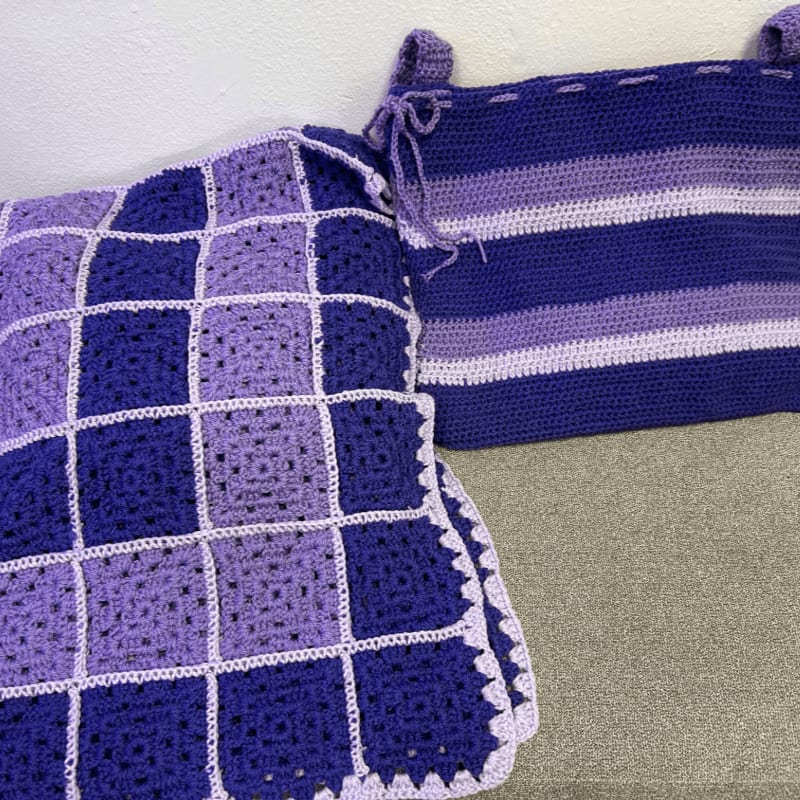 Two purple crocheted blankets, one checkered and one striped, rest on a grey surface against a plain white wall.