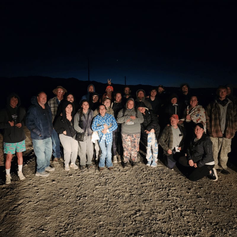 A group of people pose together on a sandy ground against a dark night sky and distant mountains.