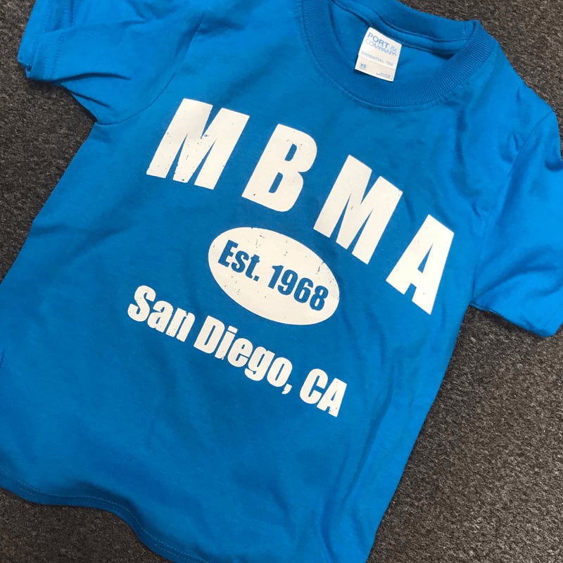 A blue t-shirt with white text reading "MBMA Est. 1968 San Diego, CA" lies on a dark grey textured surface.
