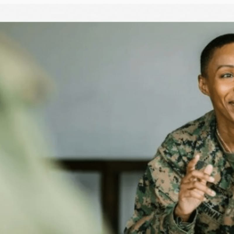 A person in military camouflage is smiling and gesturing in the foreground against a blurred, neutral-toned background.