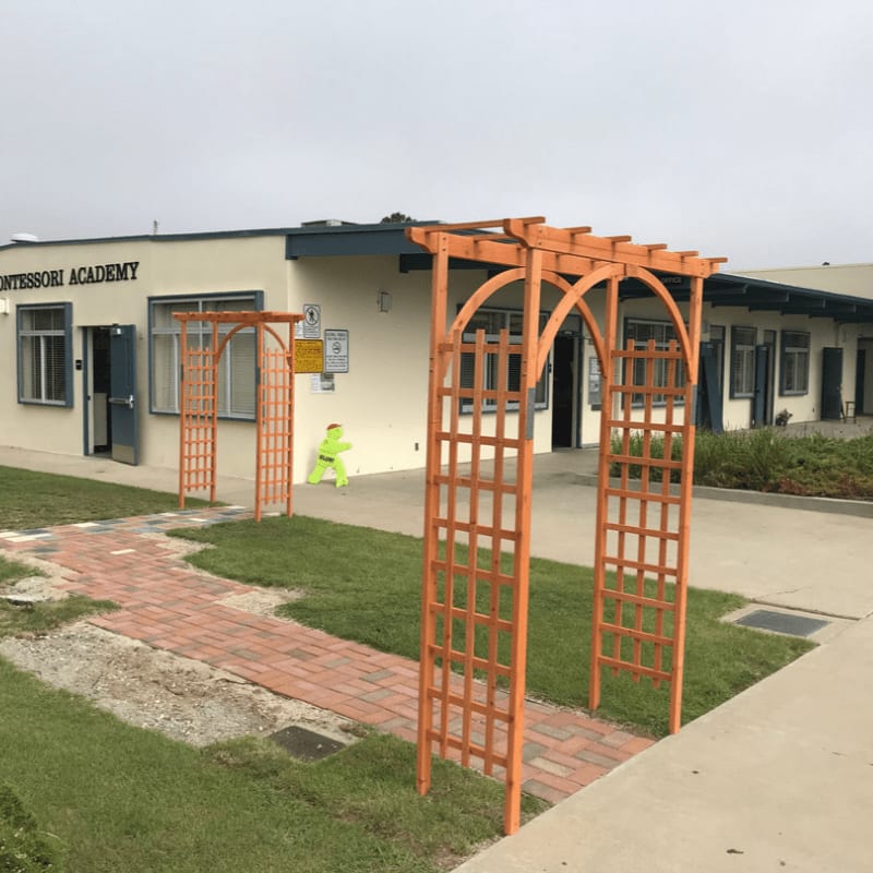 Two orange wooden trellises stand over a brick path in the foreground, with a Montessori Academy building and a small yellow figure in the background.