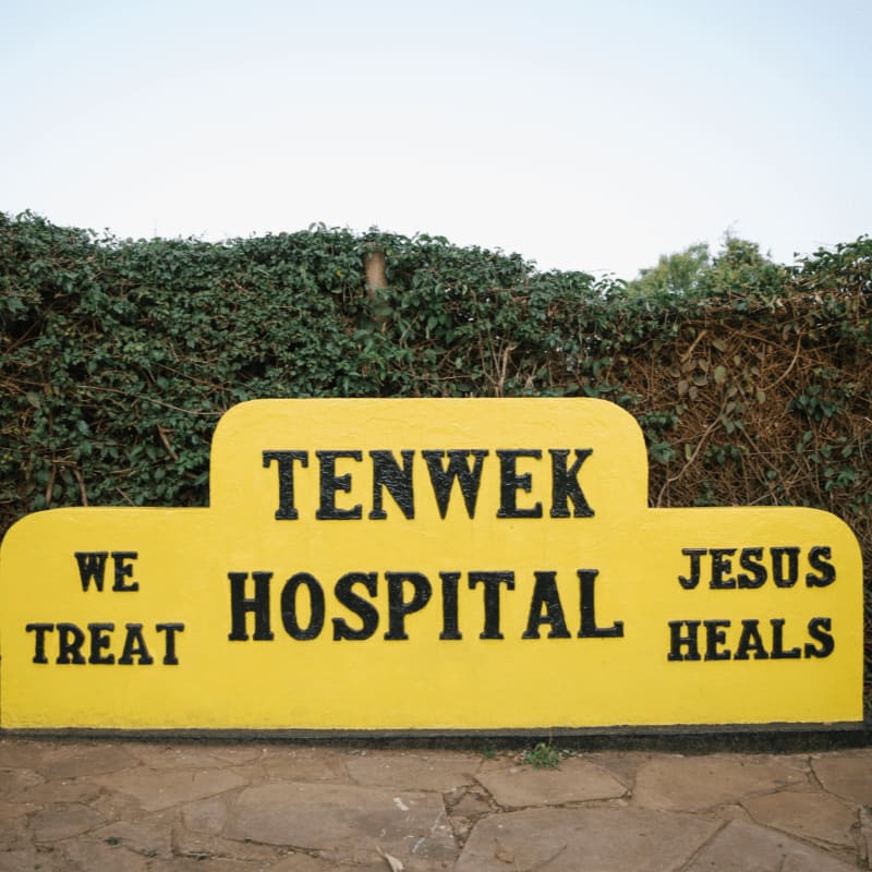 A yellow sign reading "TENWEK HOSPITAL" with the phrases "WE TREAT" and "JESUS HEALS" sits in the foreground against a background of dense green hedges.