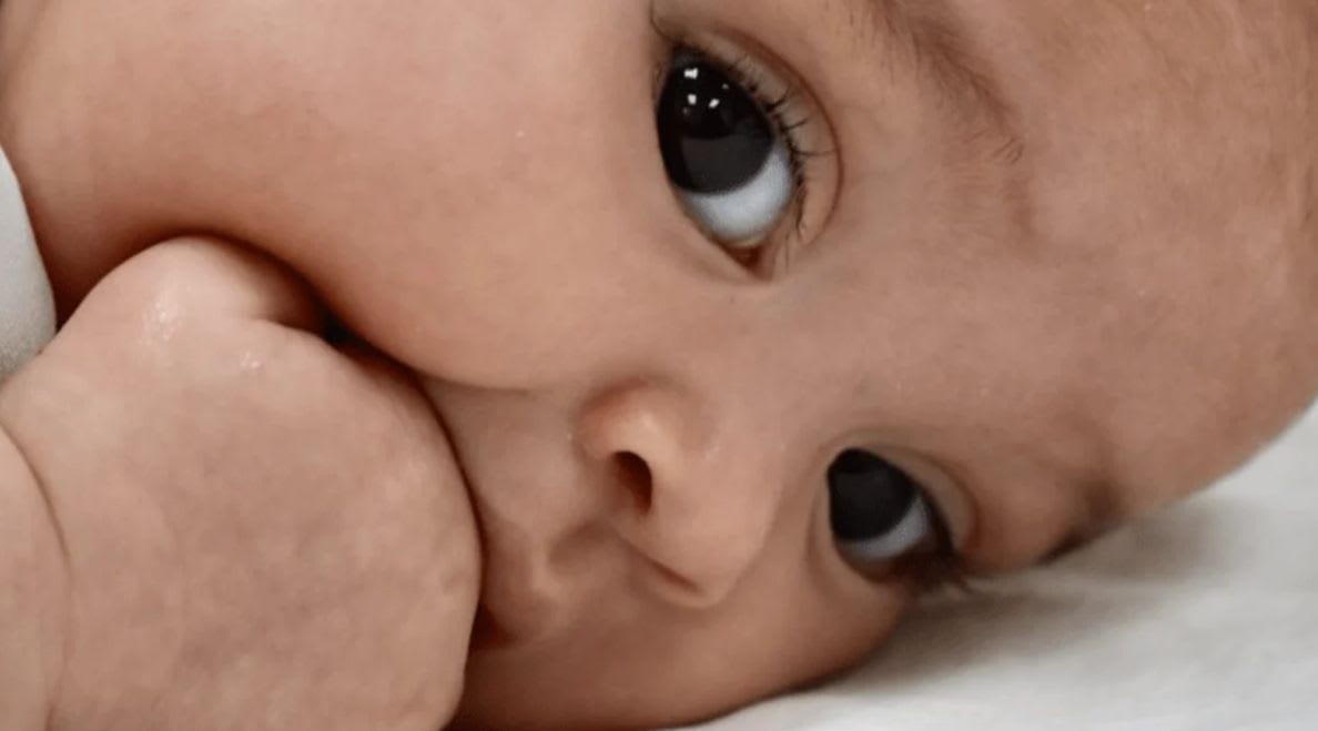 A close-up of a baby's face with a hand near its mouth against a soft, white background.