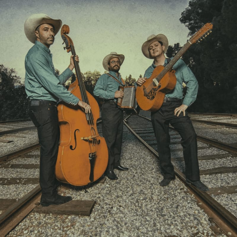 Three men in cowboy hats and blue shirts hold a double bass, an accordion, and a guitar while standing on railroad tracks against a backdrop of distant trees.