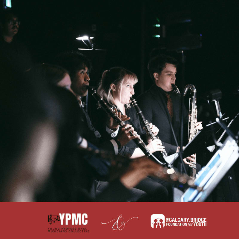 Musicians play clarinets and a bassoon in a dimly lit concert hall with a dark background.