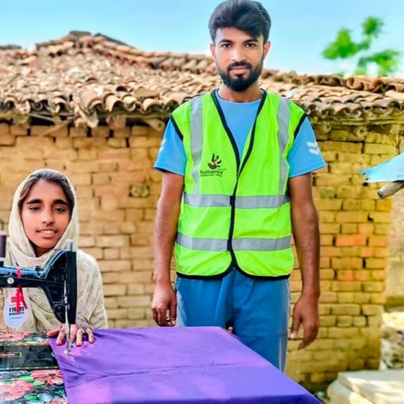 A woman sits at a sewing machine with purple fabric in the foreground, while a man in a neon yellow safety vest stands beside her against a rustic brick building.