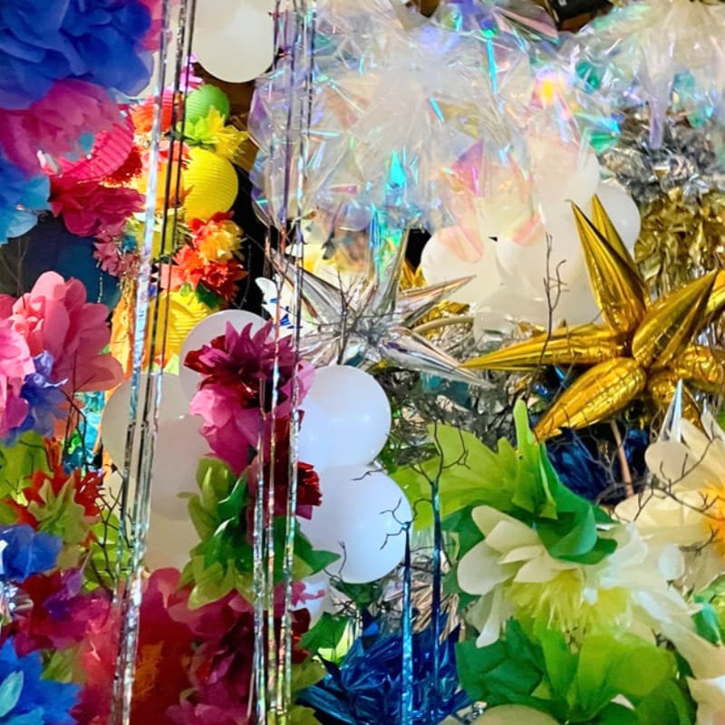 A vibrant explosion of colorful tissue paper flowers and metallic streamers fills the foreground, while a shimmering, iridescent background hints at more festive decorations.